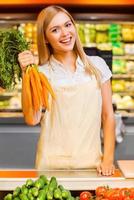 vitamine per il clienti. allegro giovane femmina venditore sorridente a telecamera e Tenere carote mentre in piedi a un' cibo memorizzare foto