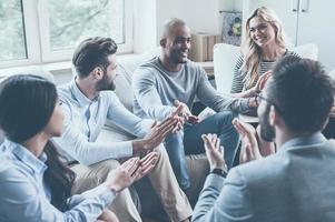 io fatto esso gruppo di giovane allegro persone seduta nel cerchio e Applaudire mentre uno uomo Esprimere a gesti e sorridente foto