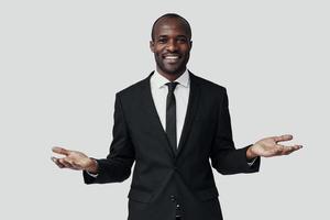 elegante giovane africano uomo nel vestito formale guardare a telecamera e sorridente mentre in piedi contro grigio sfondo foto