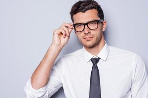 fiducioso e riuscito. ritratto di bello giovane uomo nel camicia e cravatta regolazione il suo occhiali e guardare a telecamera mentre in piedi contro grigio sfondo foto