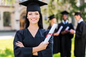 contento per essere laureato. contento giovane donna nel la laurea abiti Tenere diploma e sorridente mentre sua amici in piedi nel il sfondo foto