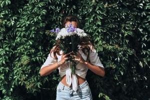 appena essere se stessa. avvicinamento di attraente giovane donna nel casuale indossare copertura viso con un' mazzo di fiori mentre in piedi nel davanti di il edera all'aperto foto