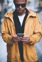 digitando Messaggio per amico. bello giovane uomo Tenere inteligente Telefono e guardare a esso mentre in piedi su il città strada foto