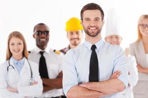 quando io crescere su io volontà essere un' uomo d'affari. fiducioso giovane uomo nel camicia e cravatta conservazione braccia attraversato e sorridente mentre gruppo di persone nel diverso professioni in piedi nel il sfondo foto