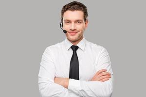 maschio operatore. bello giovane uomo nel vestito formale e cuffia guardare a telecamera e sorridente mentre in piedi contro grigio sfondo foto