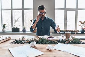 soggiorno collegato. bello giovane uomo parlando su inteligente Telefono e accartocciando carta mentre seduta nel il ufficio foto