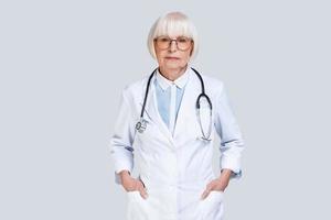 affidabile medico. bellissimo anziano donna nel laboratorio cappotto guardare a telecamera mentre in piedi contro grigio sfondo foto