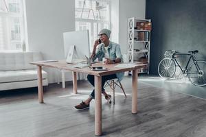 piacevole parlare. bello giovane uomo nel occhiali parlando su inteligente Telefono e sorridente mentre seduta a il suo Lavorando posto nel creativo ufficio foto