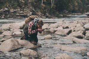 bello giovane moderno uomo potabile acqua a partire dal il fiume mentre escursioni a piedi nel montagne foto