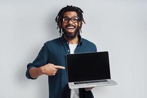 affascinante giovane africano uomo nel occhiali puntamento copia spazio su il computer portatile mentre in piedi contro grigio sfondo foto