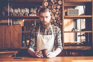 fiducioso barista. giovane barbuto uomo nel grembiule guardare a telecamera e Tenere caffè tazza mentre in piedi a bar contatore foto