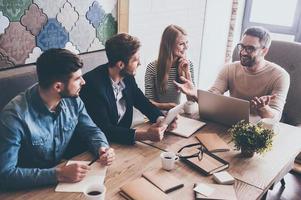 così che cosa noi dovrebbero fare Il prossimo superiore Visualizza di giovane bello uomo nel bicchieri Esprimere a gesti e discutere qualcosa con il suo collaboratori con Sorridi mentre seduta a il ufficio tavolo foto