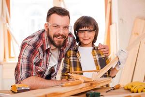 carpenteria è un' famiglia attività commerciale. allegro giovane maschio falegname Abbracciare una persona il suo figlio mentre pendente a il di legno tavolo con diverso Lavorando utensili posa su esso foto