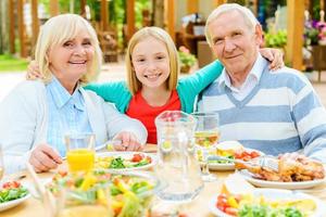 godendo tempo con nonni. contento poco ragazza Abbracciare una persona sua nonni e guardare a telecamera mentre seduta a il cenare tavolo all'aperto insieme foto