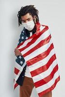 bello giovane africano uomo coperto con americano bandiera indossare medico viso maschera e guardare a telecamera mentre in piedi contro grigio sfondo foto
