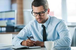 uomo d'affari a opera. fiducioso giovane uomo d'affari nel vestito formale scrittura nel Nota tampone mentre seduta a il suo Lavorando posto foto