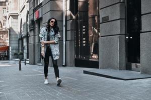 godendo strada stile. pieno lunghezza di giovane attraente donna regolazione sua manica mentre a piedi giù il strada foto