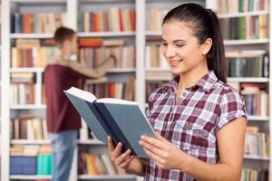 studenti nel biblioteca. bellissimo giovane donna lettura un' libro mentre uomo la scelta libro a partire dal il libro mensola su il sfondo foto