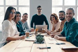 giovane e inteligente. gruppo di giovane moderno persone nel inteligente casuale guardare a telecamera e sorridente mentre Lavorando nel il creativo ufficio foto