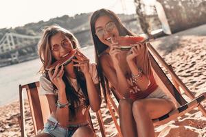 momenti di felicità. Due attraente giovane donne sorridente e mangiare anguria mentre seduta su il spiaggia foto