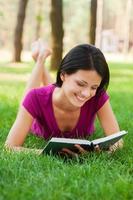 alunno nel natura. attraente giovane donna lettura libro e sorridente mentre dire bugie nel erba foto