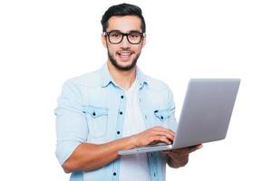 sempre pronto per aiuto. fiducioso giovane indiano uomo Tenere il computer portatile e sorridente mentre in piedi contro bianca sfondo foto