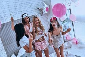 gioia di sospeso su insieme. superiore Visualizza di quattro attraente giovane sorridente donne nel pigiama potabile Champagne mentre avendo un' sonno festa nel il Camera da letto foto