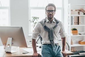 fiducioso uomo. bello giovane uomo nel casuale Abiti guardare a telecamera mentre pendente su il scrivania a il suo Lavorando posto nel creativo ufficio foto