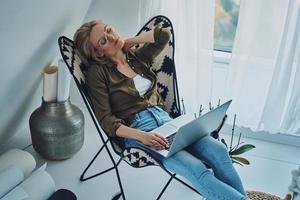 stanco giovane donna trasporto il computer portatile e conservazione occhi chiuso mentre seduta nel confortevole sedia a casa foto