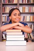 lei gli amori studiando. fiducioso giovane nero donna pendente a il libro pila e sorridente mentre seduta a il biblioteca scrivania foto