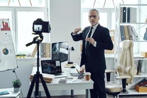 anziano uomo nel elegante attività commerciale completo da uomo condivisione il suo attività commerciale Esperienza mentre fabbricazione sociale media video foto