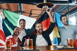 contento giovane uomini coperto nel internazionale bandiere potabile birra e Guardando sport gioco mentre seduta nel il pub foto