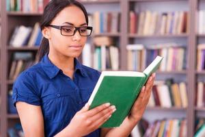 concentrato su lettura. fiducioso africano femmina alunno lettura un' libro mentre in piedi nel biblioteca foto