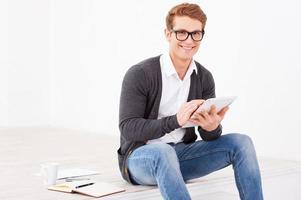 Lavorando con piacere. bello giovane uomo Lavorando su digitale tavoletta e sorridente mentre seduta su il pavimento con Nota tampone e tazza di caffè vicino lui foto