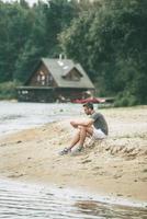 ispirando con musica e fresco aria. bello giovane uomo nel gli sport capi di abbigliamento indossare cuffie e guardare lontano mentre seduta su il Riva del fiume foto