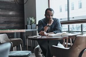 raccolta pertinente informazione. riflessivo giovane uomo conservazione mano su mento e guardare a il libro mentre seduta a il suo Lavorando posto nel il caffetteria foto