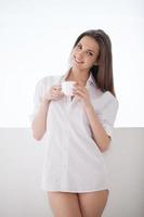 potabile sua mattina caffè. bellissimo giovane donna nel bianca camicia e mutandine Tenere un' caffè tazza e sorridente mentre in piedi vicino finestra davanzale foto