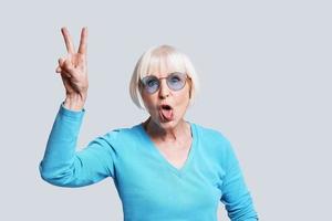 pazzo signora. giocoso anziano donna fabbricazione pace cartello e fabbricazione un' viso mentre in piedi contro grigio sfondo foto