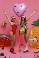 semplicemente contento. pieno lunghezza di Due attraente giovane donne nel costumi da bagno sorridente e Esprimere a gesti mentre in posa contro rosa sfondo foto