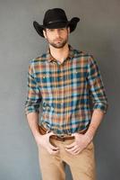 coraggioso cowboy. bello giovane uomo indossare cowboy cappello e guardare a telecamera mentre in piedi contro grigio sfondo foto