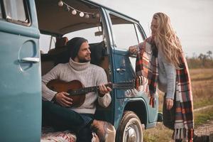 ascoltando per il canzone di amore. bello giovane uomo giocando chitarra per il suo bellissimo fidanzata mentre seduta nel blu retrò stile mini furgone foto