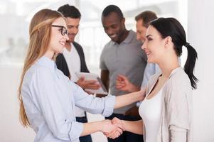 donne tremante mani. Due bellissimo giovane donne stretta di mano mentre gruppo di persone comunicare su sfondo foto