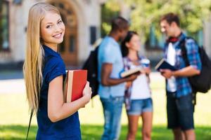 mio futuro è nel mio mani. bellissimo giovane donna Tenere libri e sorridente mentre in piedi vicino Università edificio e con sua amici chat nel il sfondo foto