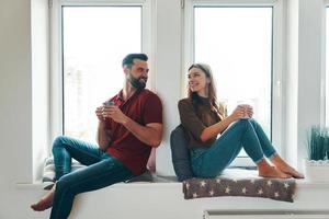 spensierato giovane coppia nel casuale capi di abbigliamento bonding insieme e sorridente mentre seduta su il finestra davanzale in casa foto