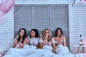 appena essere te stesso tra amici quattro attraente giovane donne nel pigiama potabile cocktail mentre dire bugie nel il letto foto