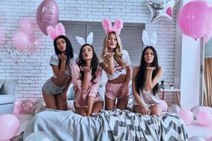 catturare quattro giocoso giovane sorridente donne nel coniglietto orecchie soffiaggio un' bacio mentre seduta su il letto con palloncini in giro foto