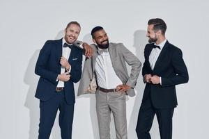 tre bello giovane uomini nel tute e Cravatte guardare a telecamera e sorridente mentre in piedi contro grigio sfondo foto