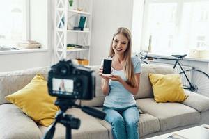 bellissimo giovane donna sorridente e puntamento copia spazio su sua inteligente Telefono mentre fabbricazione sociale media video a casa foto