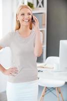 nel toccare con il colleghi. bellissimo giovane donna parlando su il mobile Telefono e sorridente mentre in piedi vicino sua Lavorando posto nel ufficio foto