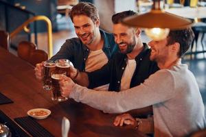 superiore Visualizza di contento giovane uomini nel casuale capi di abbigliamento tostatura ogni altro con birra e sorridente mentre seduta a il bar contatore nel pub foto